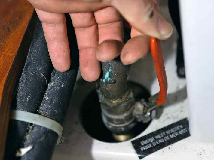 Hand inspecting an engine inlet seacock in a yacht bilge, with green verdigris corrosion visible on the fitting