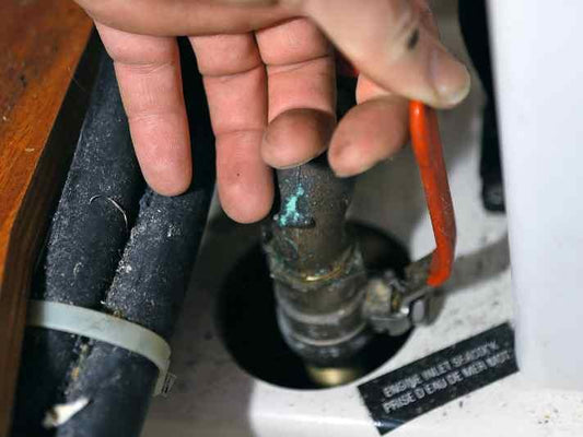 Hand inspecting an engine inlet seacock in a yacht bilge, with green verdigris corrosion visible on the fitting